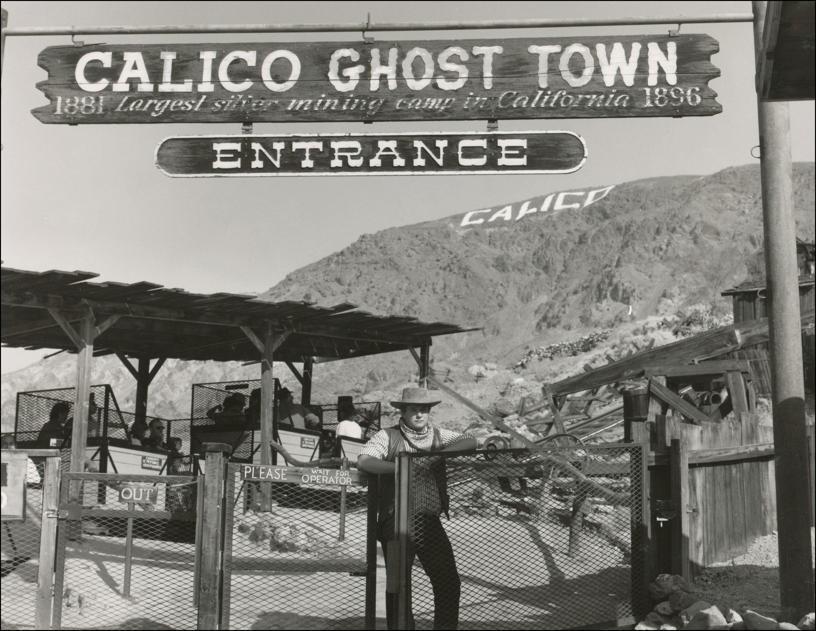 Ghost town of Calico with entrance sign. Tour trains behind cowboy standing at gate. Word Calico on the hill in the back.