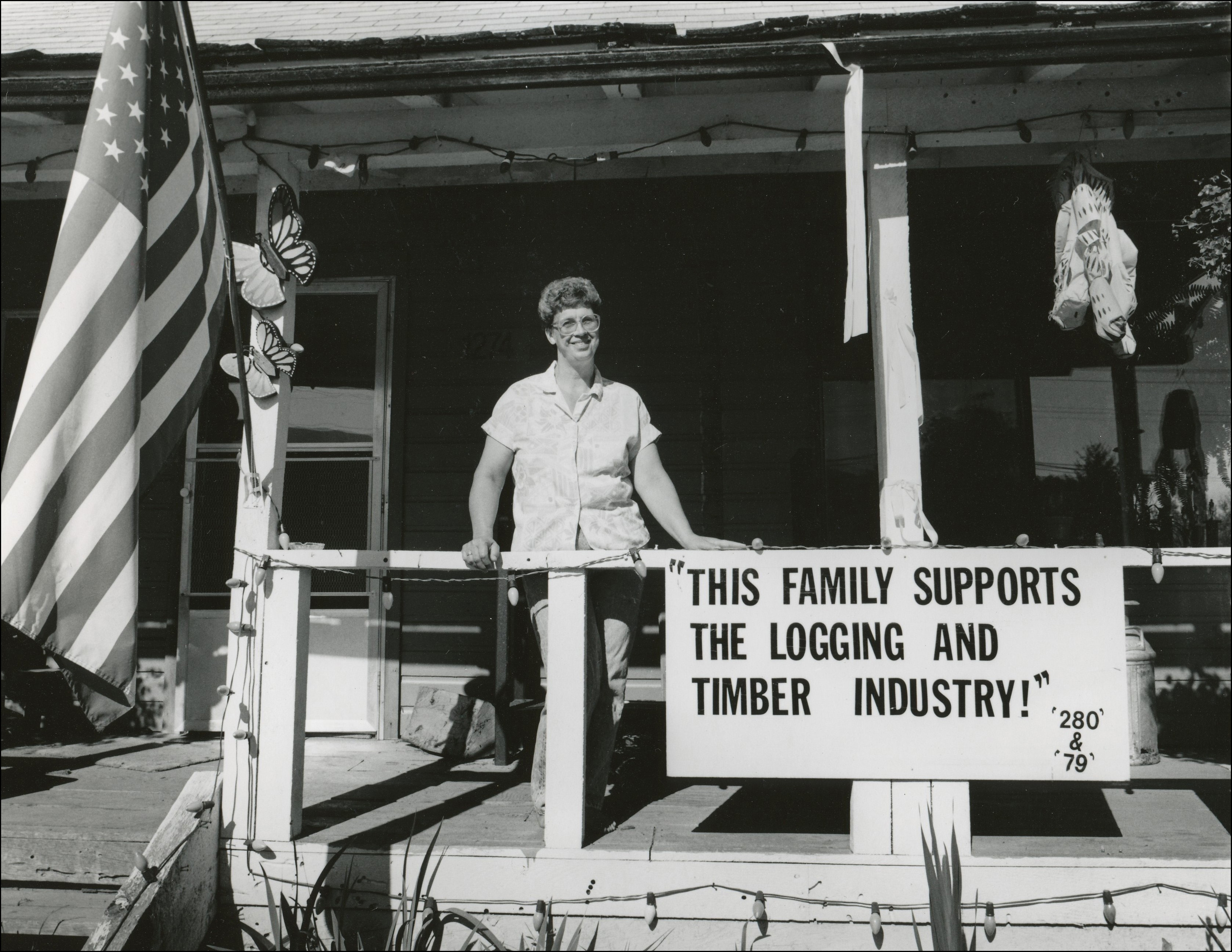 Woman standing on her front porch. American flag hanging up and a sign that says "this family supports the logging and timber industry"