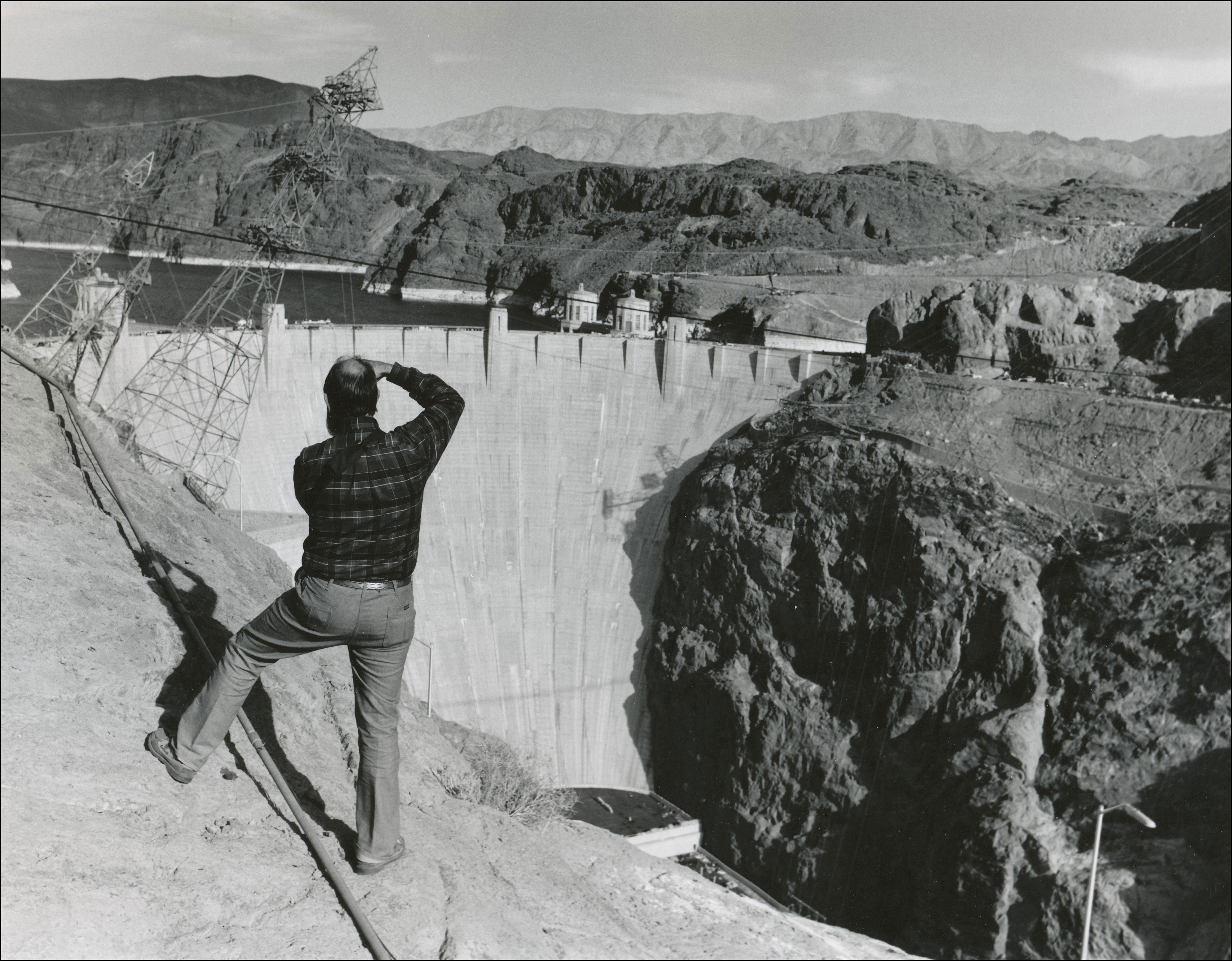 View of the back of a man standing taking a photo of hoover dam. Hoover dam in the background with water behind it.