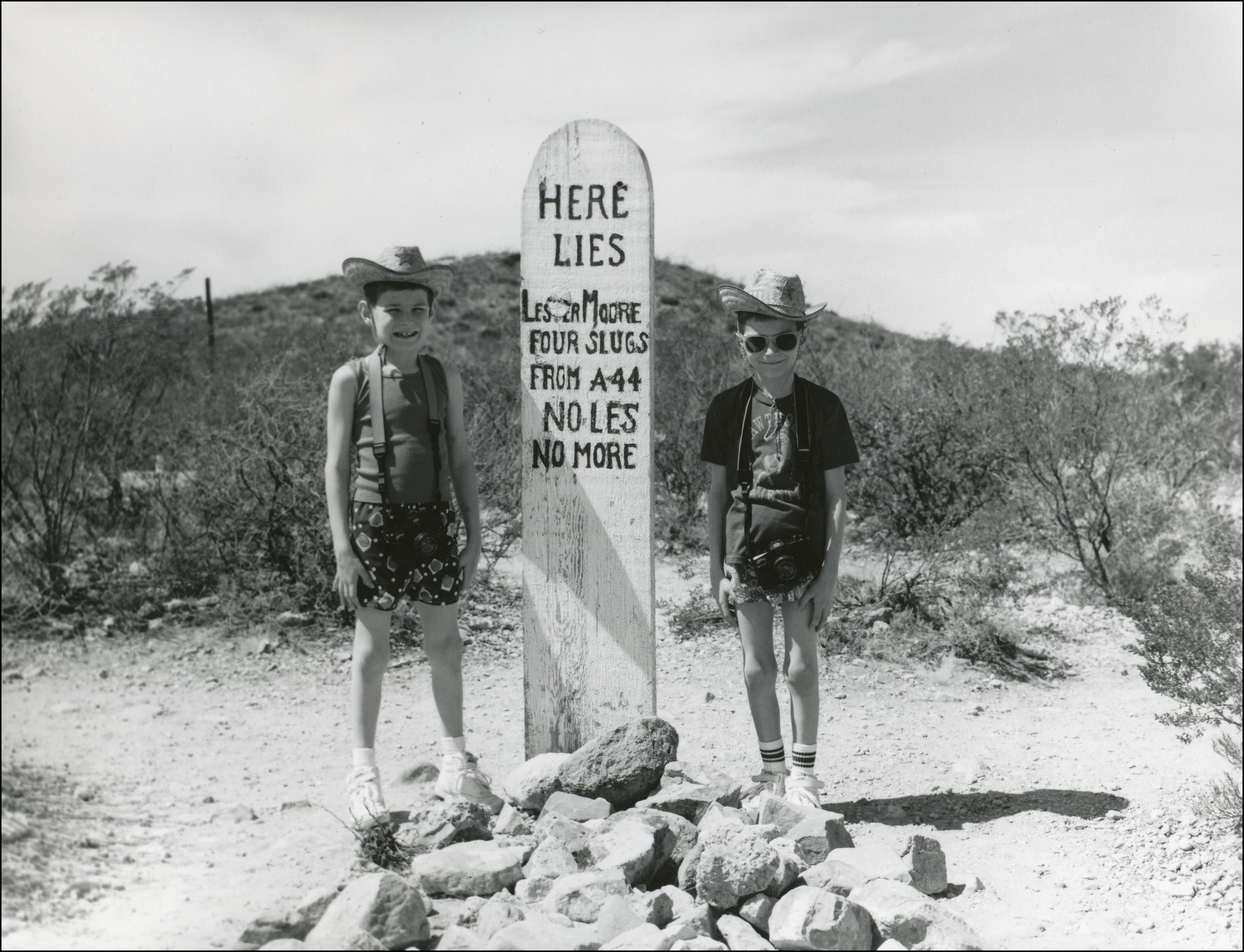 Wood marker in rock pile that says Lester Moore, Four Slugs, From A44, No Les, No More. 2 young boys, one on each side of the marker.