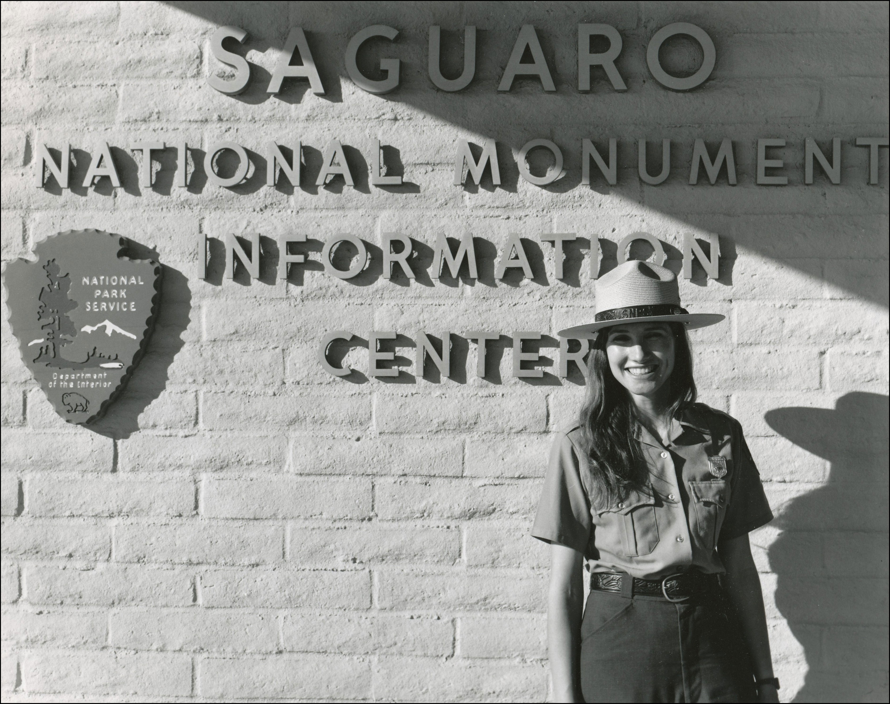 Woman Park ranger in uniform including hat standing in front of building with Naitonal Park Sign