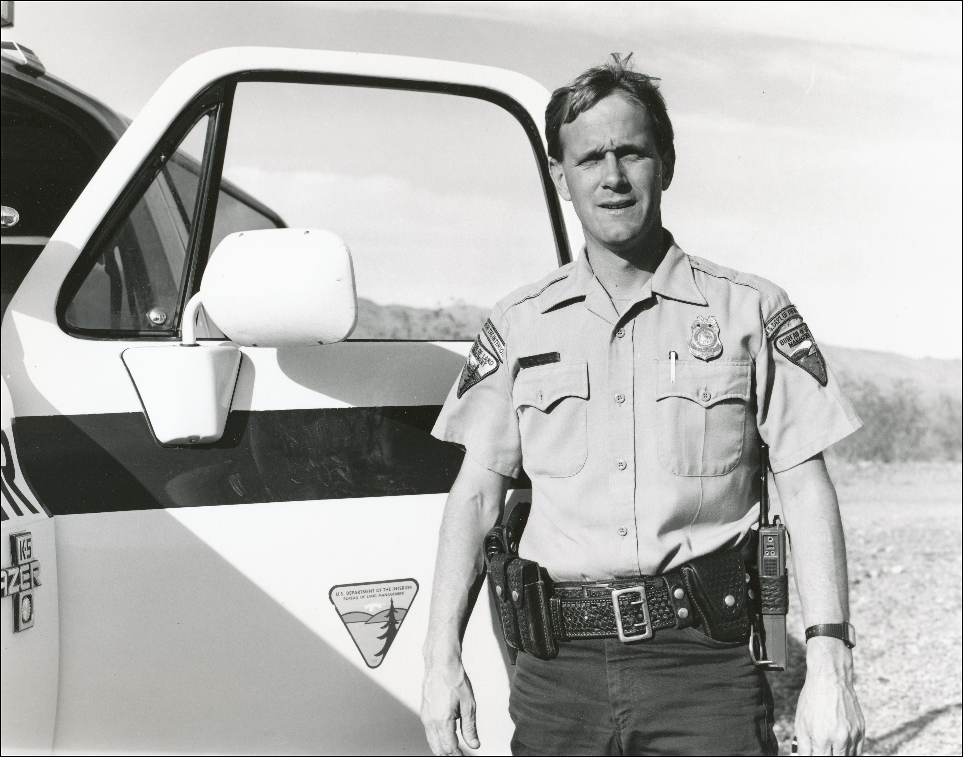 BLM officer standing in front of open driver side door of BLM truck. In uniform with badge and gun