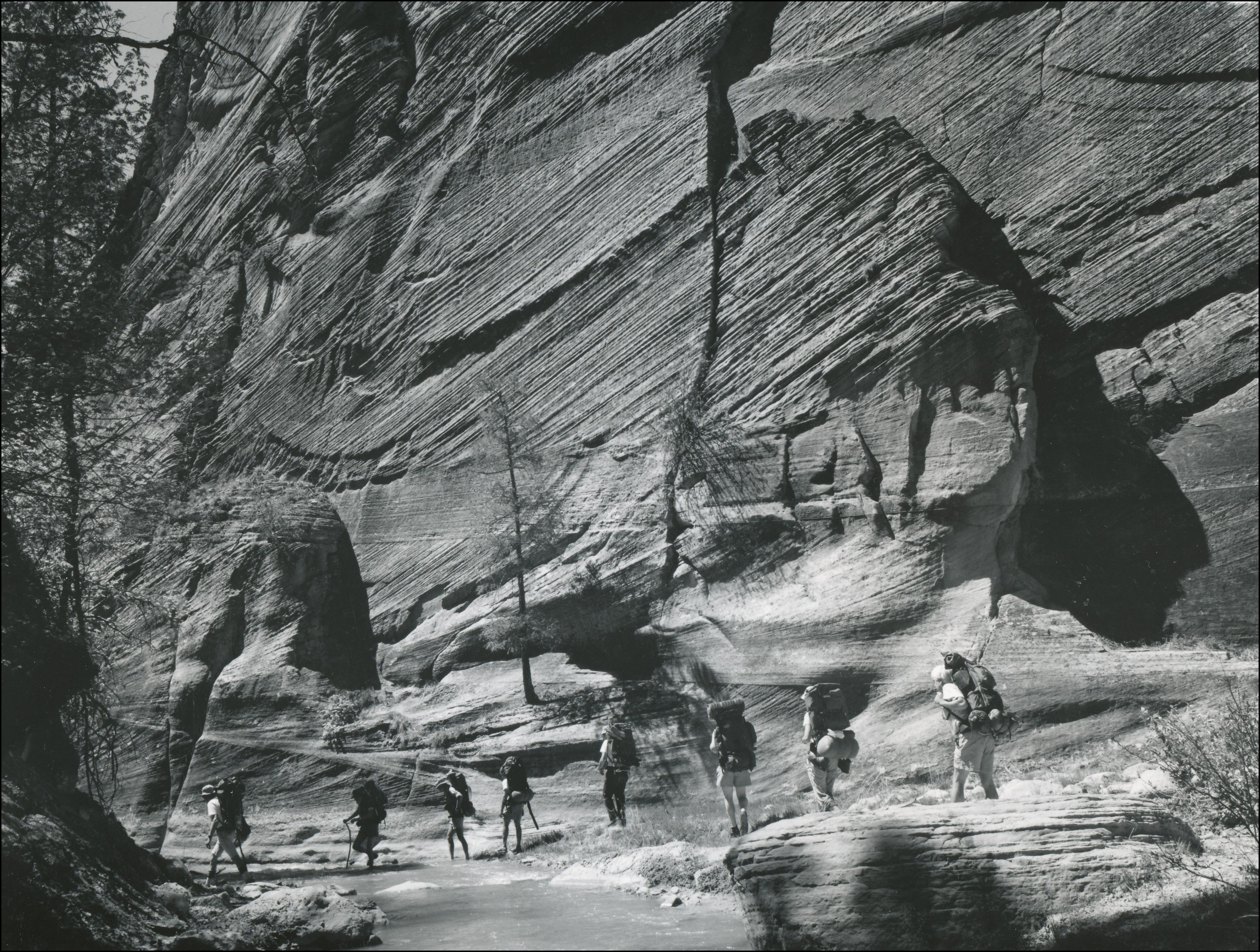 several people walking down a trail in single file at the bottom of tall rock ledges