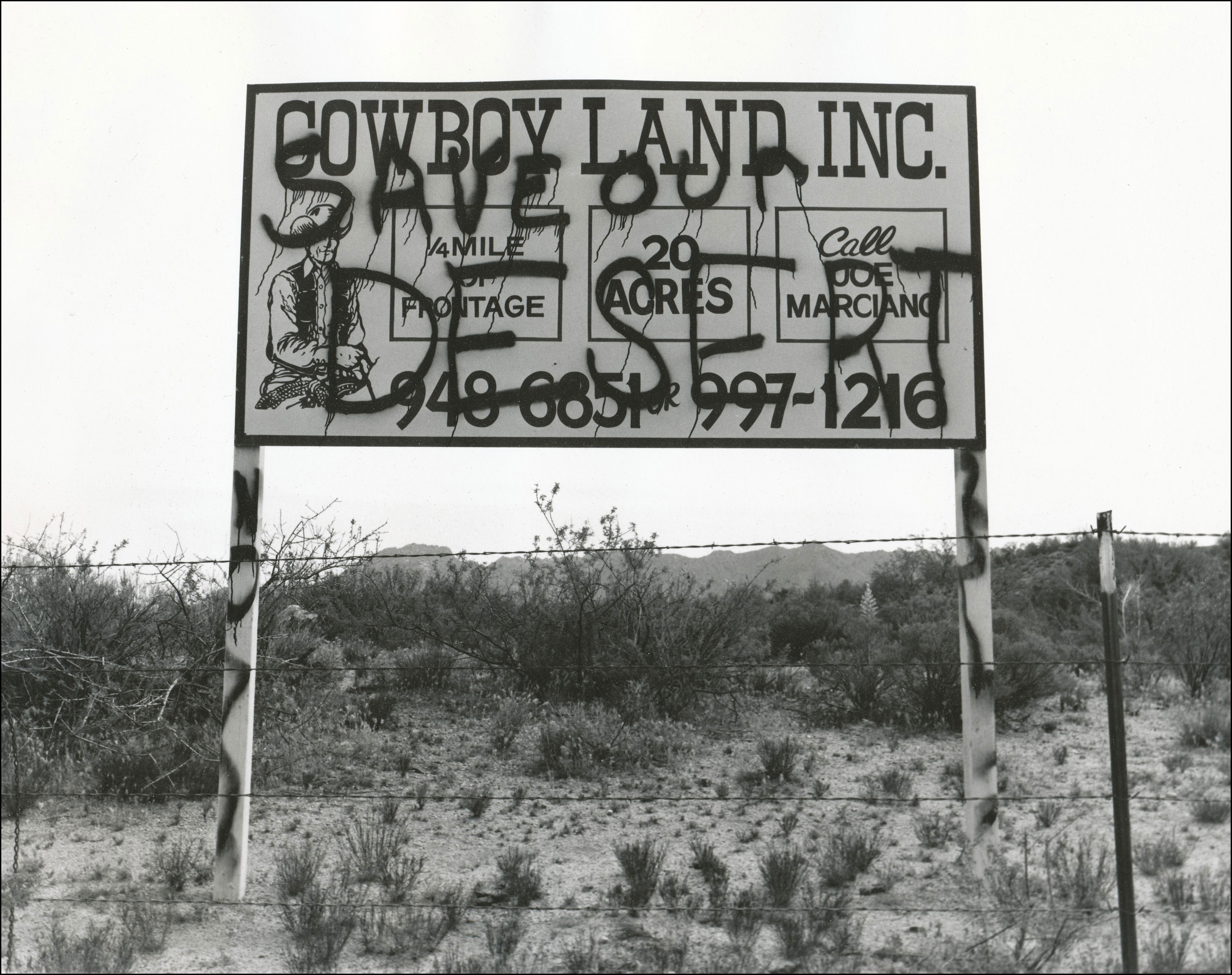 Sign on the side of a road that says Cowboy Land Inc. Quarter mile of frontage, 20 acres, call Joe Marciano, 949-6851, 997-1216. Spray painted over with the words Save our Desert