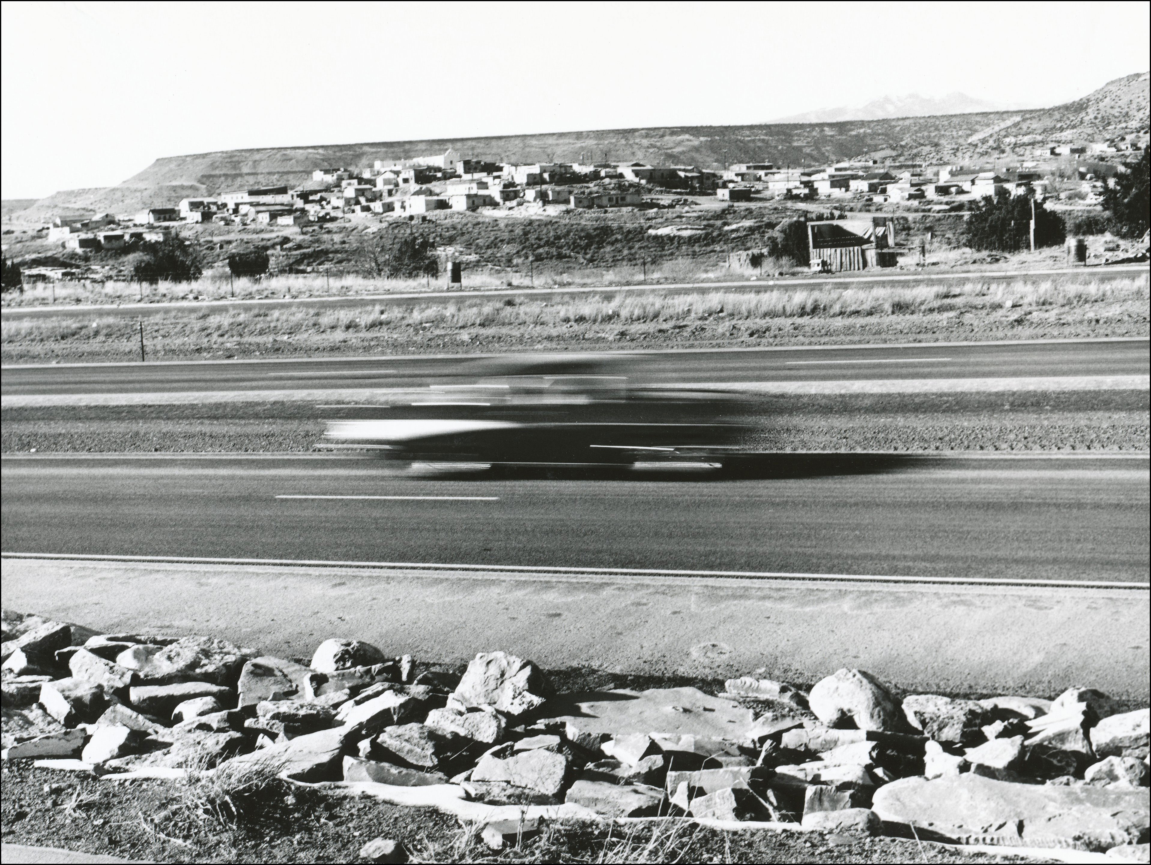 Photo of blurred car on interstate highway with a town on the otherside in the distance, on the very top of the hill is a church, a plateau is behind the town