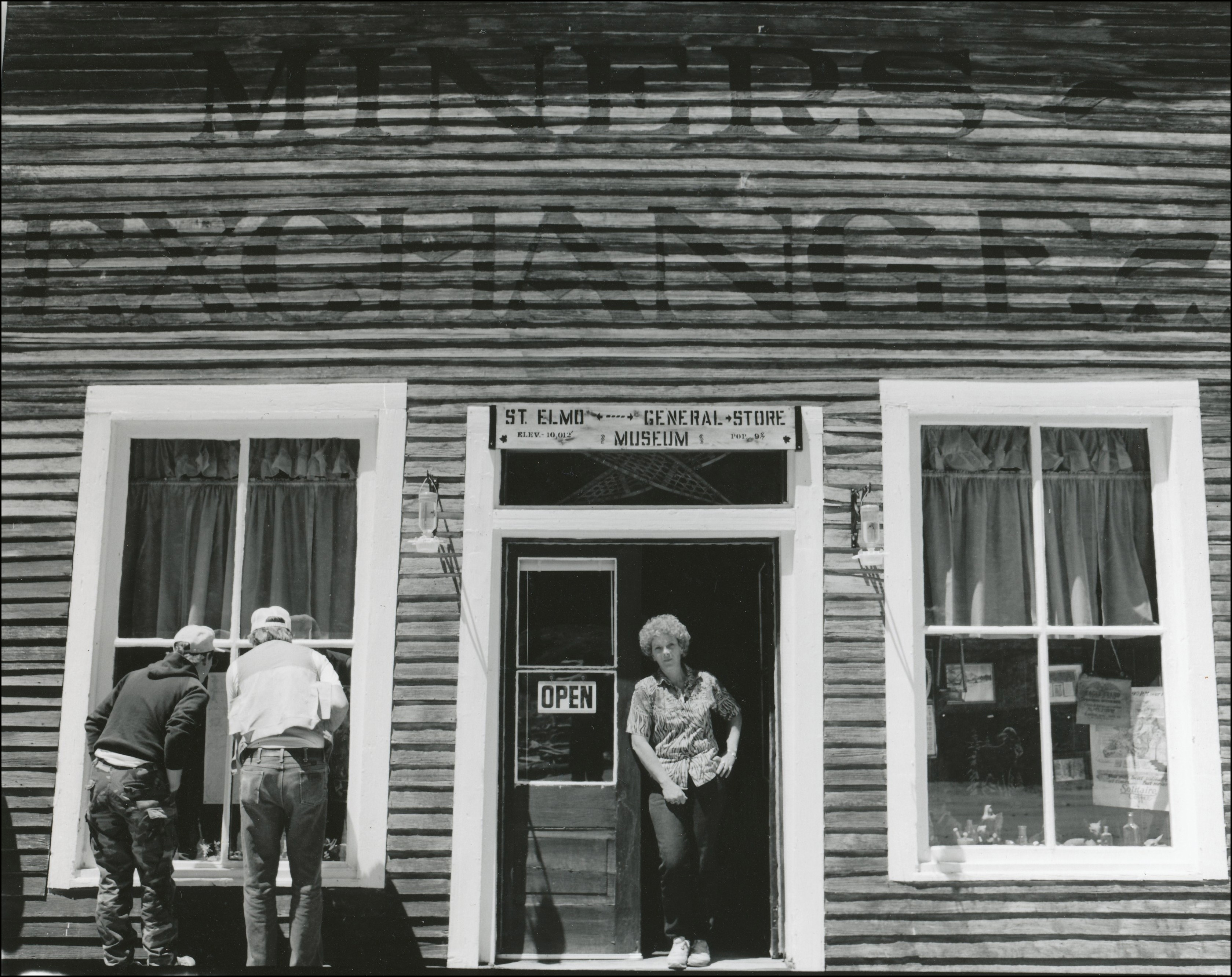 Front view of old general store with woman standing on open door and two men looking in the window