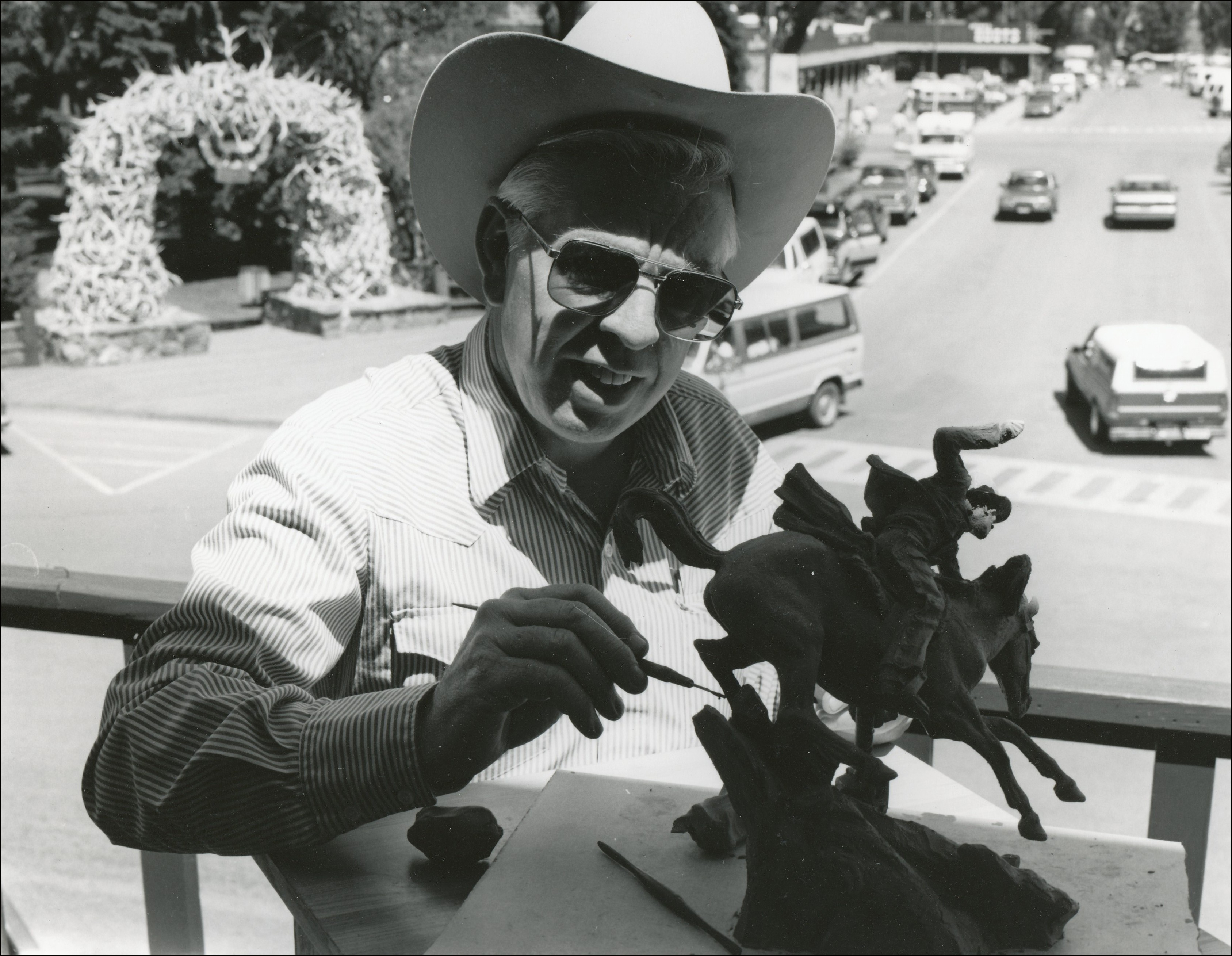 Artist wearing a cowboy hat on balcony working on a sculpture of a man running a horse down a hill