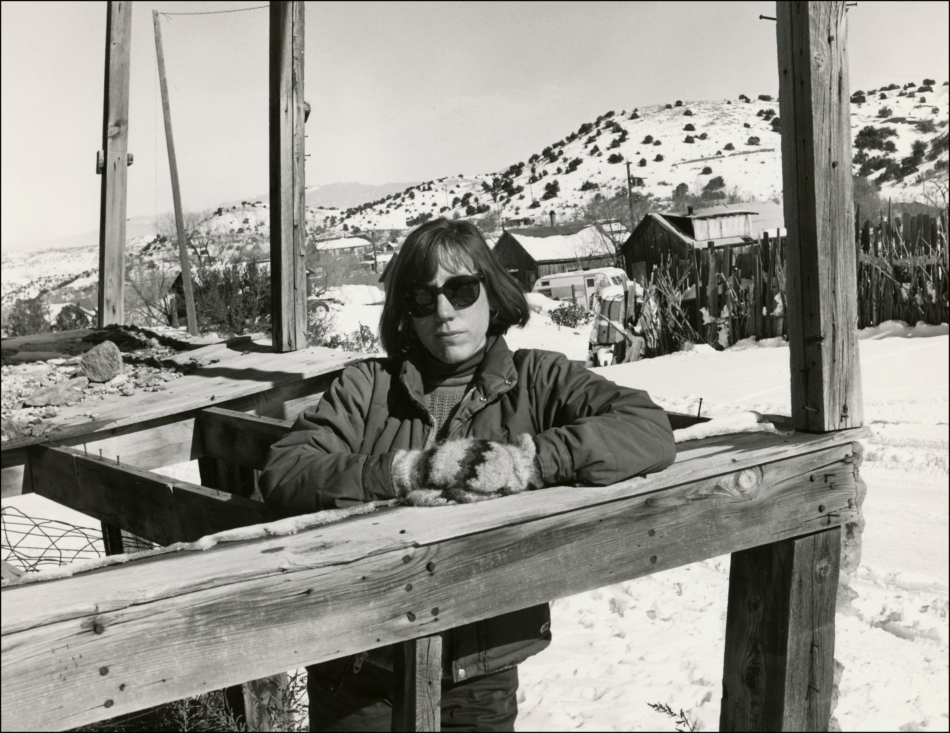 Lady with short hair and sunglasses posing with snow on the ground and some building in the background