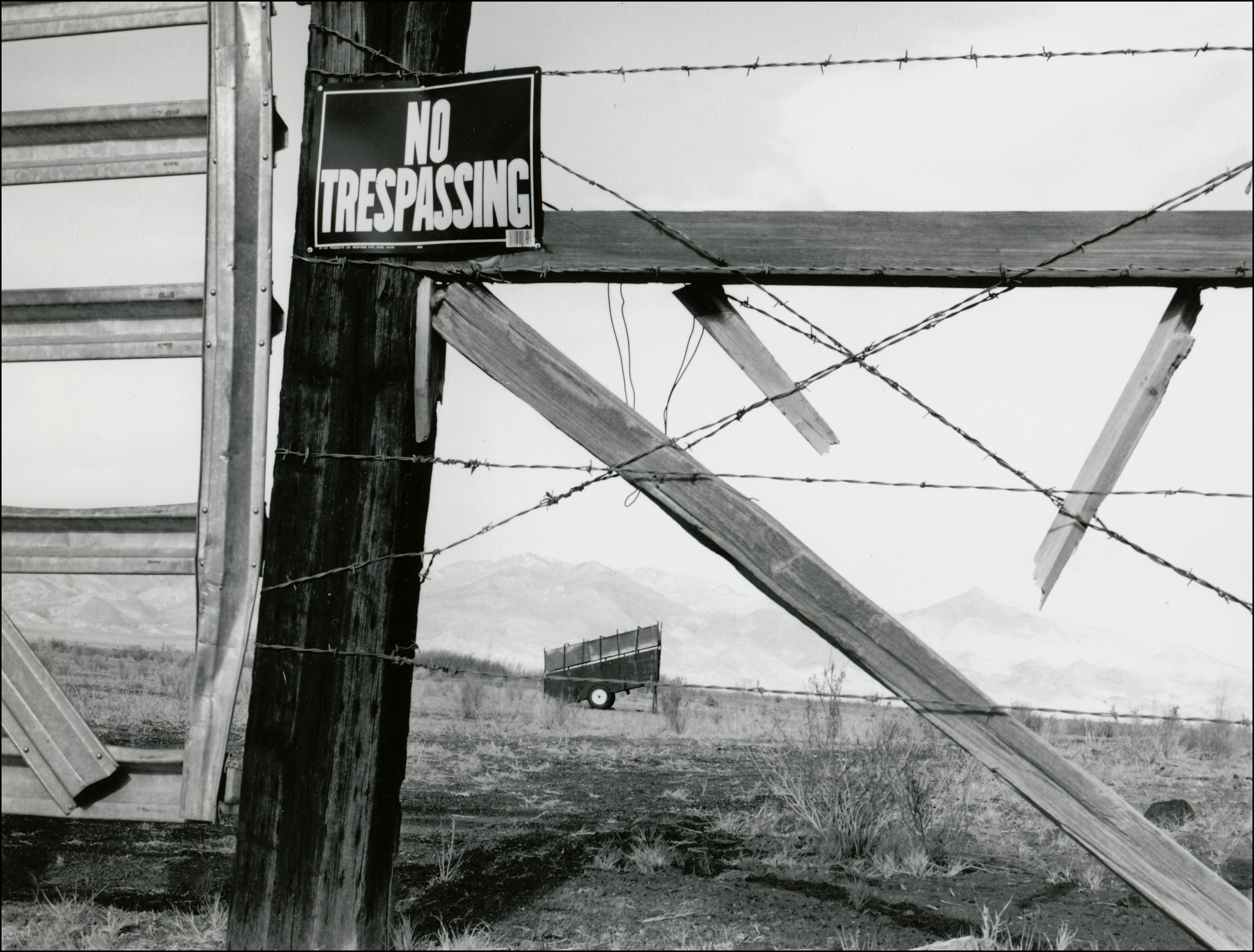Part of gate and barbed wire fence with no trespassing sign. Portable loading chute in the pasture in the background.