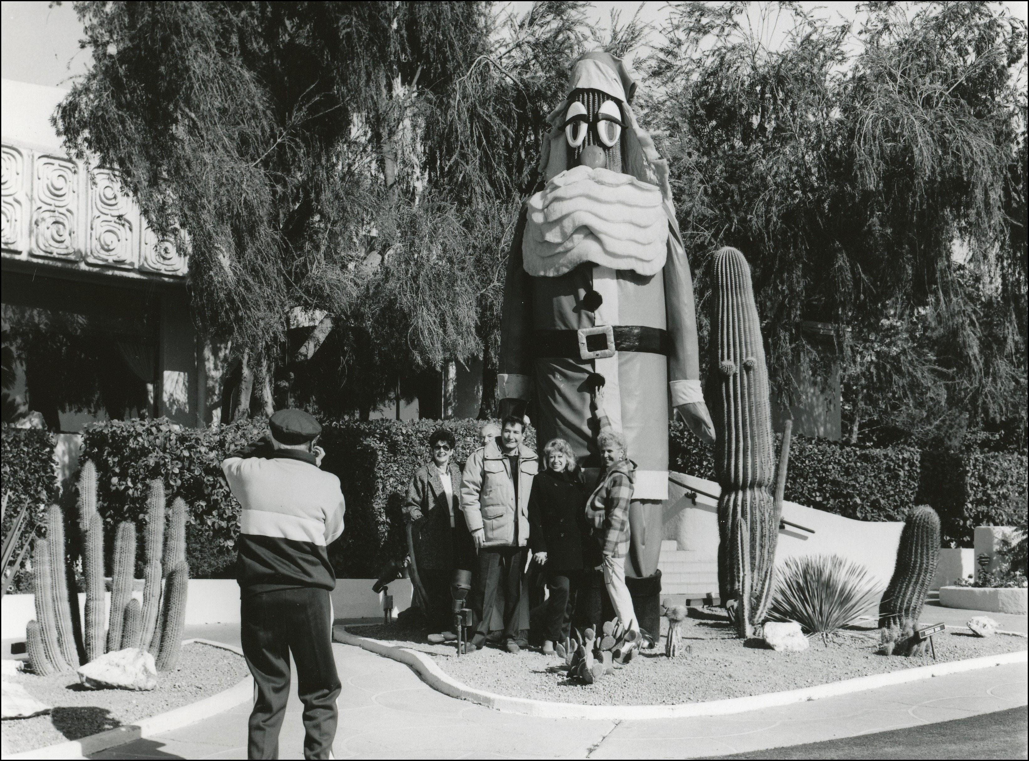 Large saguaro cactus with Santa decoration. Picture of someone taking a photo of four people standing in front of it.