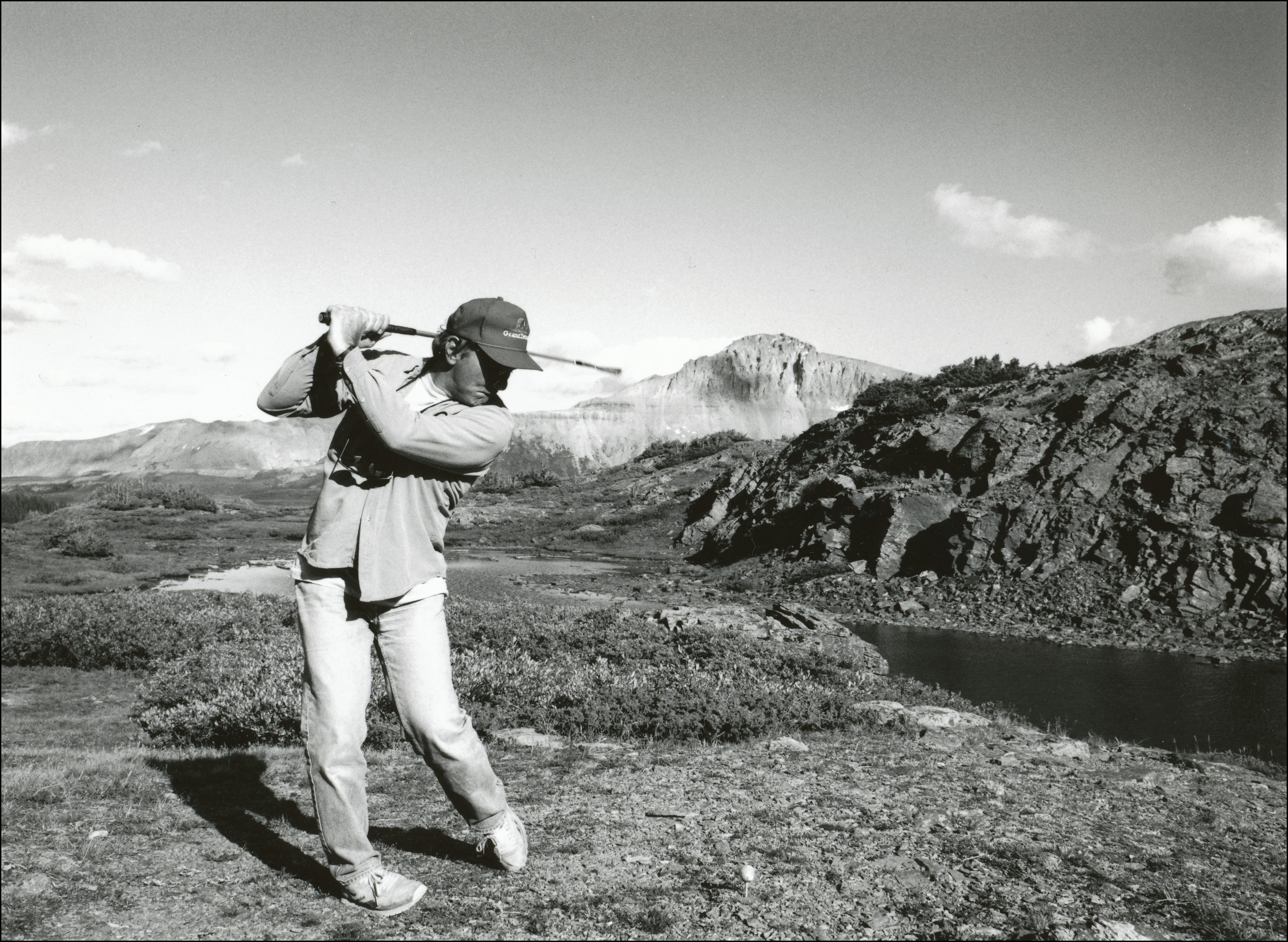 Man hitting golf ball on the edge of embankment in area with bushes, rocks and a river running in front of the man