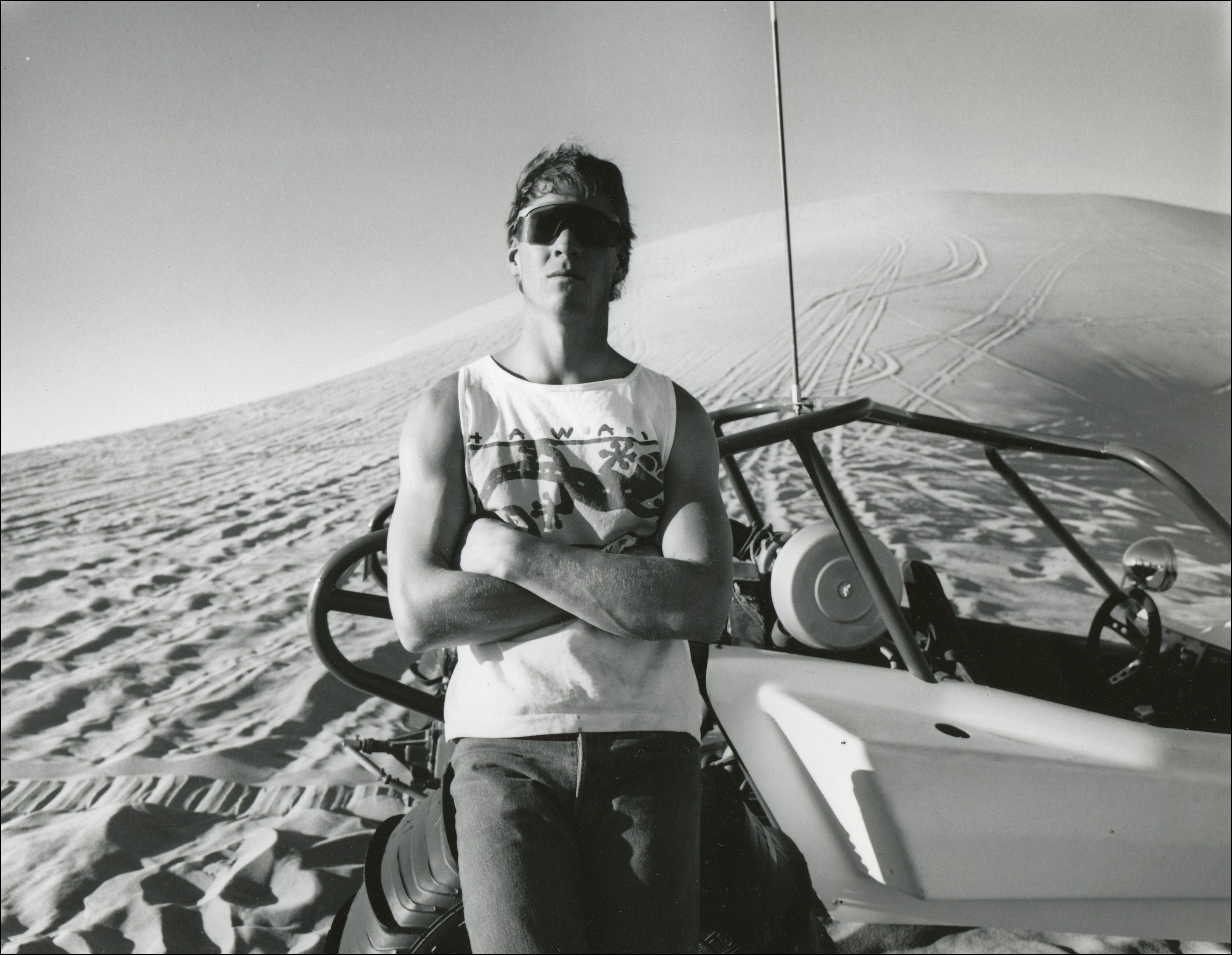 Guy in cut off sleaves and sun glasses leaning on his dune buggy in the sand