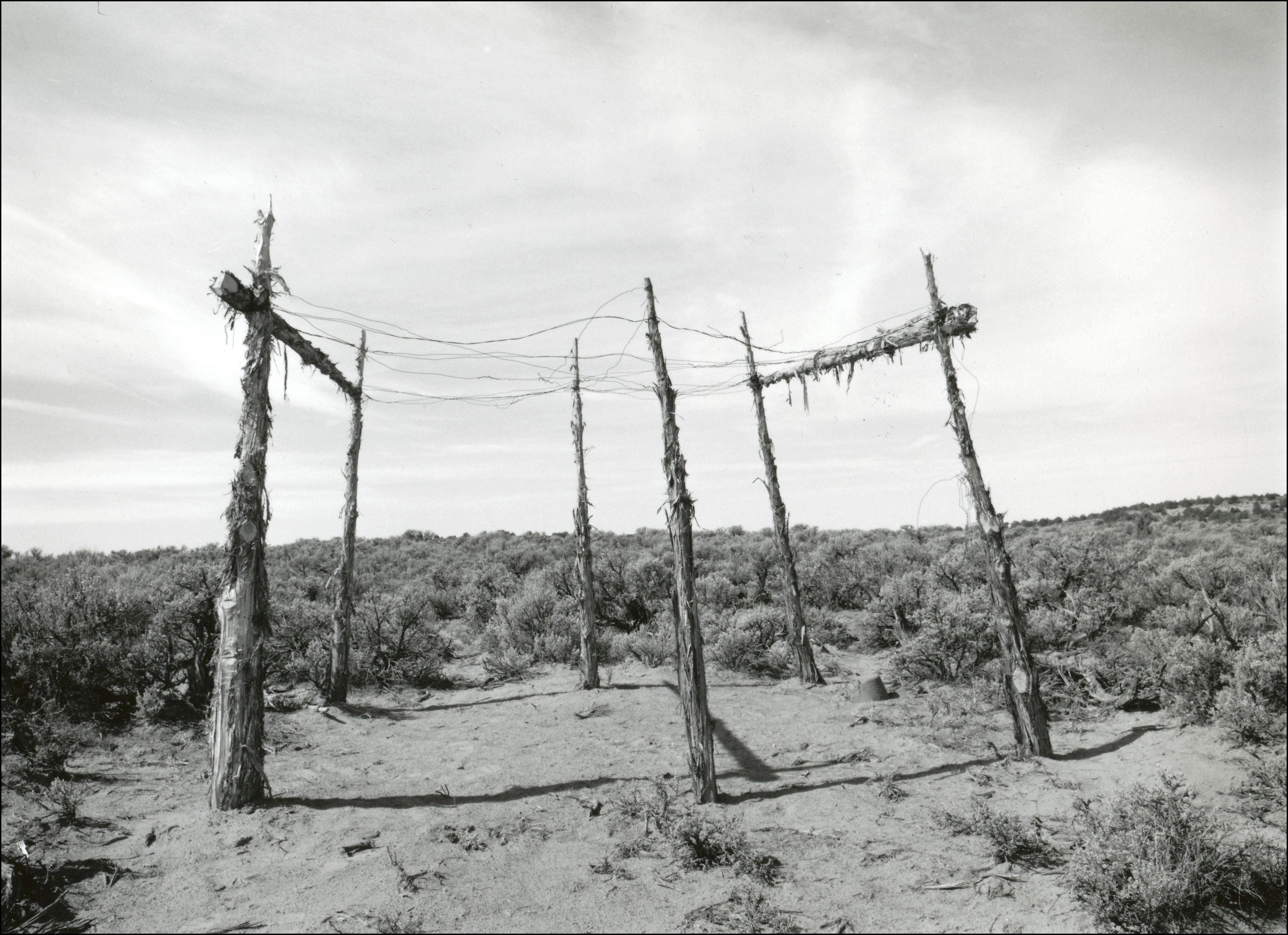 Old structure that has six cedar posts in a circle with wires on top to create some sort of roof. Dirt beneath structure and sagebrush surrounding.