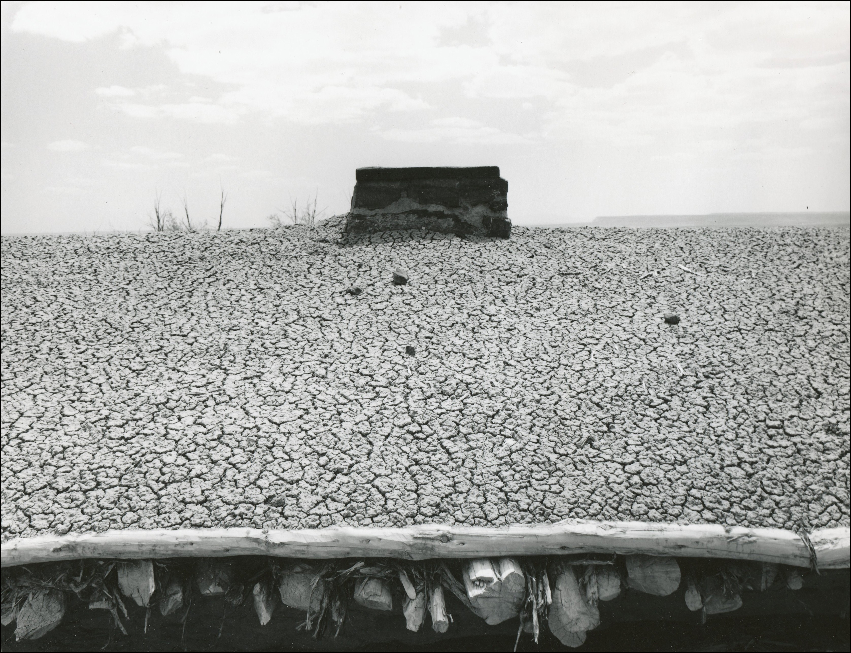 View of the roof of a home. It is covered in dirt and it's dry and cracked. If not for the wood beams in front and chimney on top would look like barren ground.