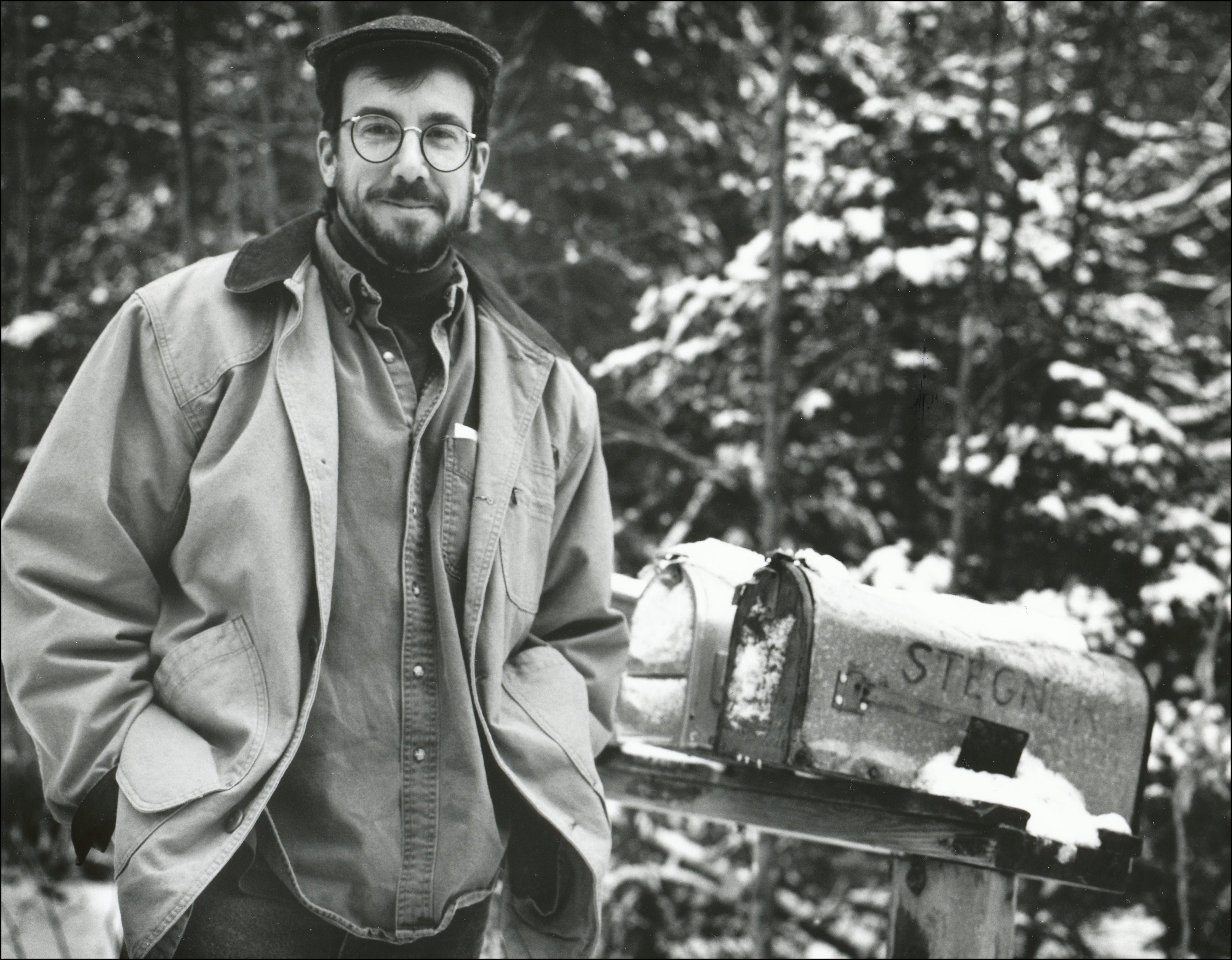 Man with hat and glasses on standing next to mailbox with the name Stegner on it. Snow on mailboxes and in pinetrees in the background